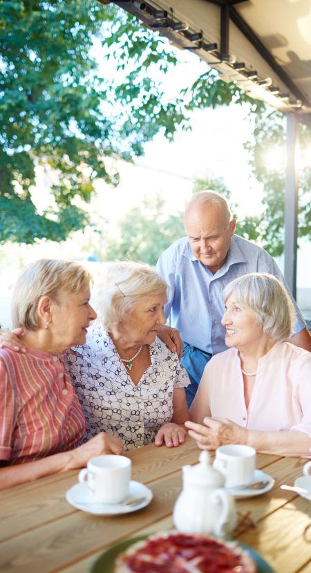 Senior friends having tea party in lovely summer cafe and sharing latest news with each other; two women putting arms on each others shoulders
